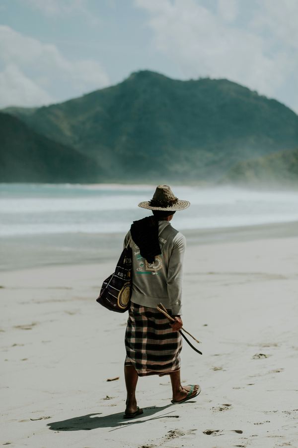 A man in a straw hat and sarong walking along a beach